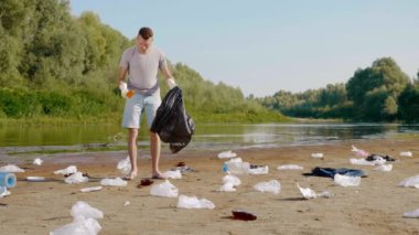 Adam kirlenmiş nehrin kıyısında plastik çöp topluyor ve baş parmağını kaldırıyor.