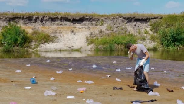 Adam kirlenmiş nehir kıyısında plastik çöp topluyor ve en sevdiği şarkıyı dinliyor. — Stok video