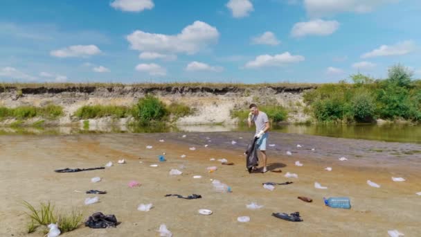 Muž sbírá plastové odpadky na březích znečištěné řeky a poslouchá oblíbenou píseň — Stock video
