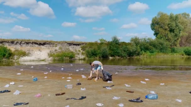 Wütender Mann sammelt Plastikmüll am Ufer des verschmutzten Flusses ein und wirft dann Müll ab — Stockvideo