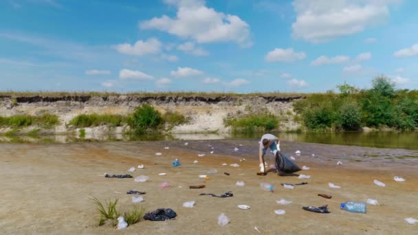 Человек собирает пластиковый мусор на берегах загрязненной реки и слушает музыку — стоковое видео