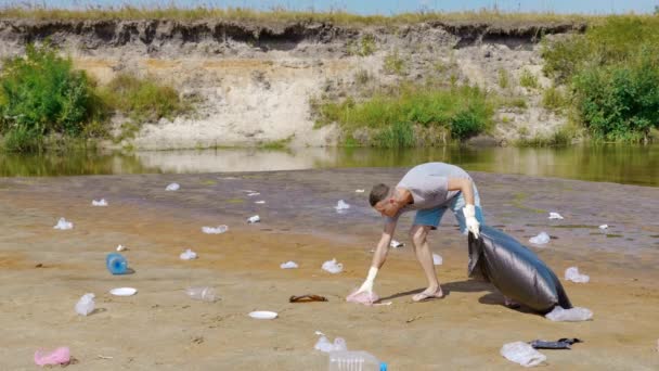 El hombre recoge basura plástica en las orillas de un río seco y contaminado — Vídeos de Stock