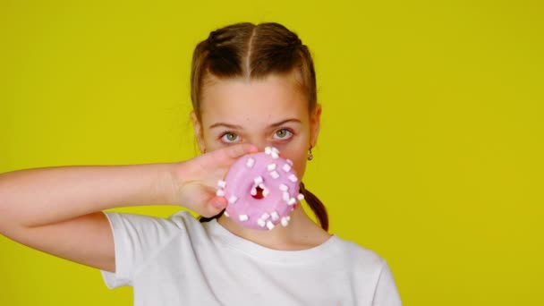 Teenage girl in a white T-shirt covers her mouth with a pink donut. — Stok video