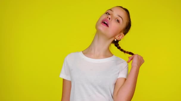 Teenage girl in a white T-shirt chews gum and twists a pigtail. — Stok video