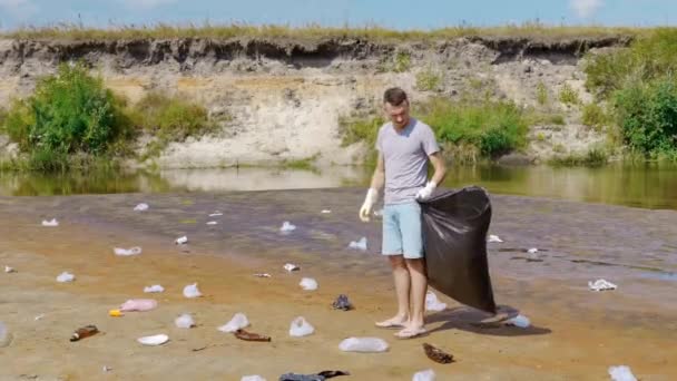 El hombre endereza la bolsa de plástico a lo largo de la basura en las orillas del río contaminado — Vídeos de Stock