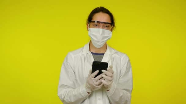 Mujer joven con abrigo médico, máscara protectora está llamando en el teléfono inteligente — Vídeo de stock