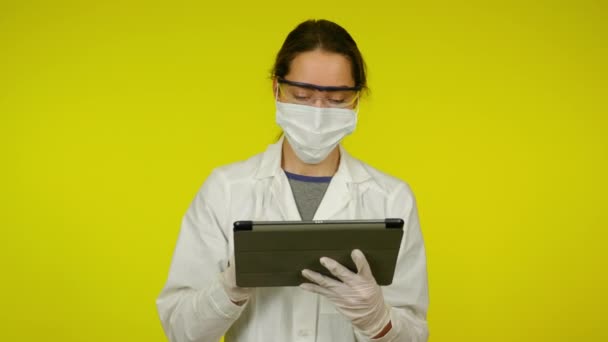 Молодая женщина в медицинском пальто, защитная маска использует таблетки — стоковое видео