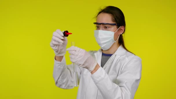 Femme en manteau médical, masque protecteur et lunettes goutte à goutte réactif dans le tube à essai — Video