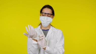 Tıbbi maskeli ve güvenlik gözlüklü genç doktor ellerine lateks eldiven takıyor.