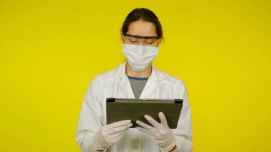 Tıbbi maskeli, gözlüklü ve lateks eldivenli genç doktor tablete bakıyor.