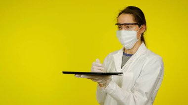 Tıbbi maskeli, gözlüklü ve lateks eldivenli doktor elinde bir tablet tutuyor.