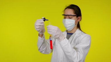 Tıp önlüklü, koruyucu maskeli ve gözlüklü kadın deney tüpüne damlatıyor.