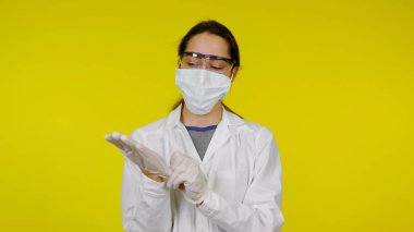Tıbbi maskeli ve güvenlik gözlüklü genç doktor ellerine lateks eldiven takıyor.