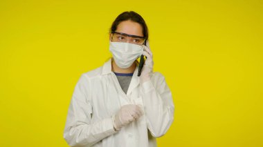 Tıbbi maskeli, gözlüklü ve lateks eldivenli genç doktor telefonda konuşuyor.