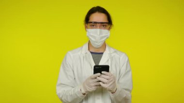 Sağlık ceketli, koruyucu maskeli genç bir kadın akıllı telefonu elinde tutuyor.