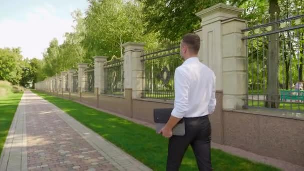 Επιχειρηματίας με ένα φορητό υπολογιστή στο χέρι περπατά μέσα από το πάρκο και κοιτάζει γύρω — Αρχείο Βίντεο