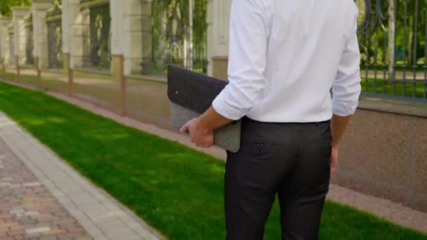 Empresario con un portátil en la mano camina por el parque y mira a su alrededor — Vídeo de stock