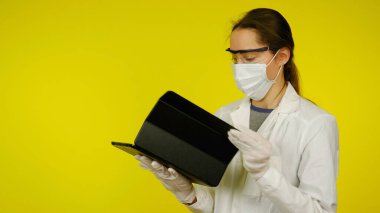 Tıbbi maskeli, gözlüklü ve lateks eldivenli genç doktor tablete bakıyor.