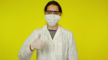 Tıp önlüklü kadın, koruyucu maske baş parmağını kaldırıyor.