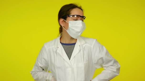 Mujer joven en abrigo médico, máscara protectora está bailando sobre fondo amarillo — Vídeos de Stock