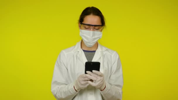 Jonge vrouw in medische jas, beschermend masker maakt gebruik van smartphone. — Stockvideo