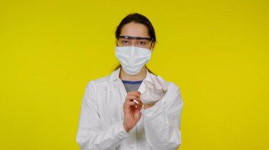 Tıbbi maskeli ve güvenlik gözlüklü genç doktor ellerine lateks eldiven takıyor.
