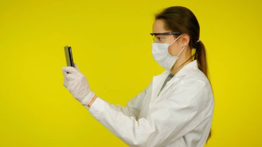 Tıbbi maskeli, gözlüklü ve lateks eldivenli genç doktor tablete bakıyor.