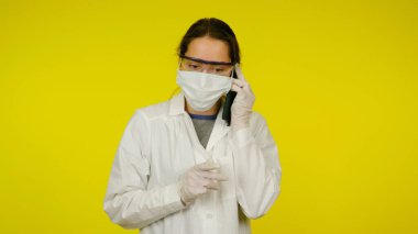Tıbbi maskeli, gözlüklü ve lateks eldivenli genç doktor telefonda konuşuyor.