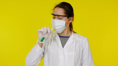 Tıbbi maskeli, gözlüklü ve lateks eldivenli doktor test tüpündeki yeşil sıvıya bakıyor.