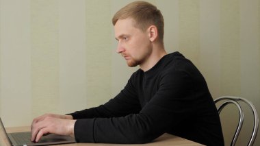 Adam masada bir sandalyeye oturur ve dizüstü bilgisayarın klavyesine metin yazar.