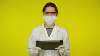 Sağlık ceketli, koruyucu maskeli genç kadın elinde bir tablet tutuyor.