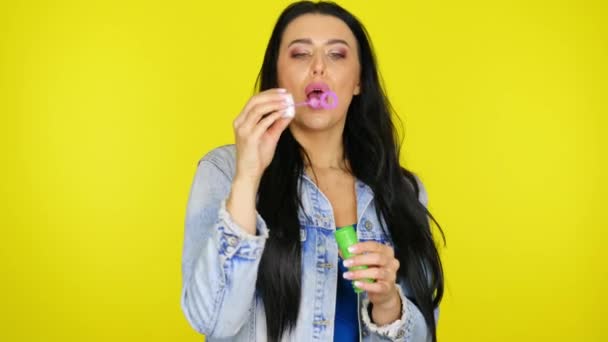 Woman inflates soap bubbles and smiles on a yellow background with copy space — Stock Video