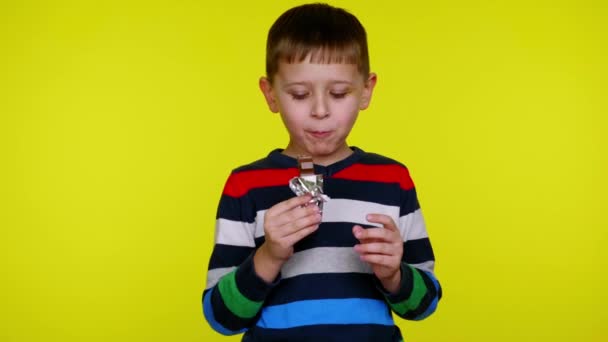 Little child boy open chocolate bar and eats on yellow background with copyspace — Stock Video