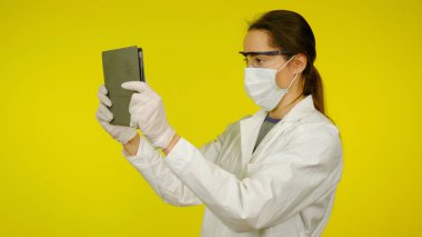 Tıbbi maskeli, gözlüklü ve lateks eldivenli genç doktor tablete bakıyor.