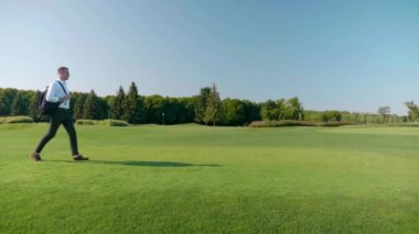 Omzunda sırt çantası olan bir iş adamı yeşil çimlerin üzerinde parkta yürüyor.