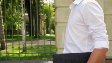 Elinde dizüstü bilgisayarı olan bir iş adamı parkta yürür ve etrafa bakar.