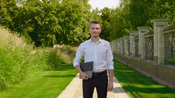 Geschäftsmann im Park mit Laptop in der Hand, in die Kamera blickend und lächelnd — Stockvideo