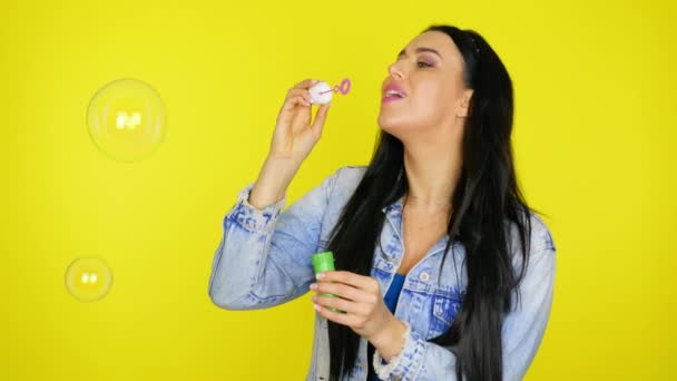 Vrouw blaast zeepbellen en glimlacht op een gele achtergrond met kopieerruimte — Stockvideo