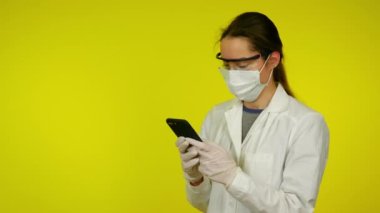 Sağlık ceketli, koruyucu maskeli genç bir kadın akıllı telefon kullanıyor.