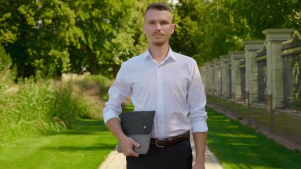 Homme d'affaires est debout dans un parc avec un ordinateur portable dans les mains, en regardant la caméra — Video