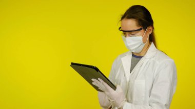 Tıbbi maskeli, gözlüklü ve lateks eldivenli genç doktor tablete bakıyor.