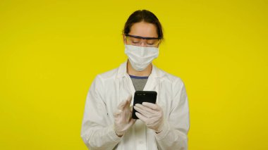 Tıbbi maskeli, gözlüklü ve lateks eldivenli doktor akıllı telefona bakar.