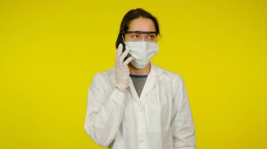 Tıbbi maskeli, gözlüklü ve lateks eldivenli genç doktor telefonda konuşuyor.