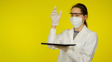 Tıbbi maskeli, gözlüklü ve lateks eldivenli doktor elinde bir tablet tutuyor.