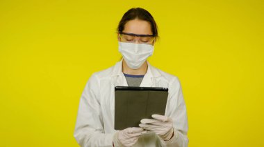 Tıbbi maskeli, gözlüklü ve lateks eldivenli genç doktor tablete bakıyor.