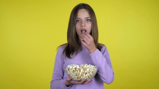 Tiener meisje eet popcorn uit een glazen kom en glimlacht op een gele achtergrond — Stockvideo