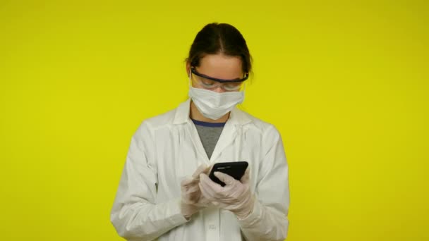 Jonge vrouw in medische jas, beschermend masker praat op smartphone — Stockvideo