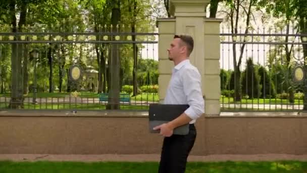 Elinde dizüstü bilgisayarı olan bir iş adamı parkta yürür ve etrafa bakar. — Stok video