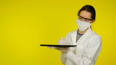 Tıbbi maskeli, gözlüklü ve lateks eldivenli doktor elinde bir tablet tutuyor.