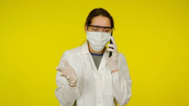 Tıbbi maskeli, gözlüklü ve lateks eldivenli genç doktor telefonda konuşuyor.